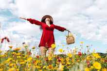 From Below Happy Female In Red Sundress, Hat And Handbag Standing With Eyes Closed On Blossoming Field With Yellow And Red Flowers With Outstretched Arms Enjoying On Warm Spring Summer Day