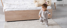 Happy Baby Boy In Grey Romper Walking On Carpet Near Soft Toy On Bed, Banner