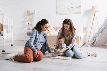 happy mother reading baby book to infant son near african american woman
