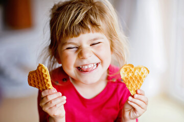 Portrait of happy little preschool girl holding fresh baked heart waffle. Smiling hungry toddler child with sweet biscuit wafer. Sweet sugar belgian waffles.