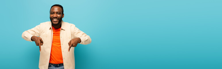 cheerful african american man pointing down with fingers isolated on blue, banner