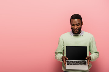 frustrated african american man showing laptop with blank screen on pink background