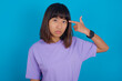 © Roquillo - Unhappy young beautiful asian woman wearing purple t-shirt against blue makes suicide gesture and imitates gun with hand, curves lips keeps two fingers on temple, shoots, being tired of everything,