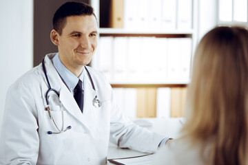 Friendly male doctor and patient woman discussing current health examination while sitting in sunny clinic. Perfect medical service in hospital