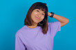 young beautiful asian woman wearing purple t-shirt against blue wall being confused and wonders about something. Holding hand on head, uncertain with doubt. Pensive concept.