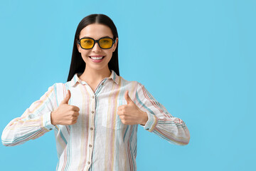 Beautiful young woman wearing stylish sunglasses and showing thumb-up on color background