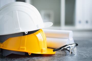 close up view of white and yellow hard safety helmet hat, blueprint, safety glasses on cement floor, suit equipment safety of engineer or worker, start project construction concept.