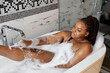 © Roman - Side view on african woman shaving her sexy legs while taking bath at home, copy space, lying in tub, young beautiful lady take care about body