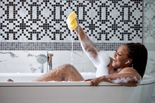 Smiling Happy African Woman Clean Fresh Healthy Black Skin Spa Treatment Enjoy Relaxing Taking Shower And Bath With Bubble Foam Spa Holding Sponge In Hands, In Bathtub At Bathroom At Home
