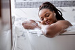 © Roman - Calm Dreamy Afro American Lady Lying In Bathtub With Foam, Relaxing In Headphones, With Eyes Closed, At Home In Bathroom