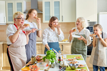 Wall Mural - Caucasian family, women of different generations enjoy eating pizza together in kitchen, after cooking time. Senior grandwomen and children standing next to table with food, wearing apron