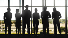 Full Length Back View Backlit Shot With Silhouettes Of Six People, Men And Women, Standing Before Panoramic Window And Looking Through Glass In Business Center Or In Airport