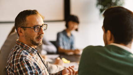 Middle aged man in eyeglasses and with beard talking to colleague sitting next to him at desk during business meeting