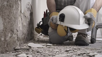 Wall Mural - house renovation concept, construction worker holds in hands helmet and safety headphones, he wears the tool bag on his belt , close up with wall plaster rubble and ladder on background