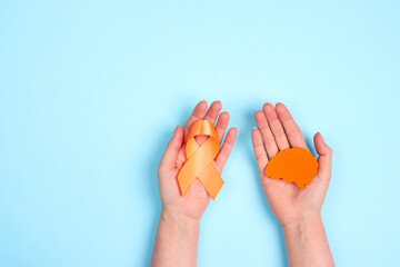 Wall Mural - Female hands hold an orange awareness ribbon and brain symbol on a blue background. World Multiple Sclerosis Day.