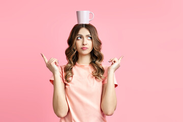 Wall Mural - Funny pretty woman having fun with cup of coffe on her head on pink background