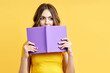 © GVS - Young woman hiding behind an open book and looking to camera isolated over yellow background