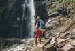 Man with backpack dressed in active trekking clothes holding trekking boots in hand near mountain river waterfall smiling and enjoying the Nature.Traveling, trekking, nature concept image