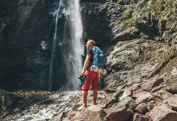 Wall Mural - man with backpack dressed in active trekking clothes holding trekking boots in hand near mountain ri