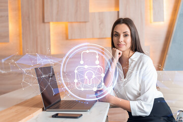 Attractive businesswoman in white shirt at workplace working with laptop to defend customer cyber security. Concept of clients information protection. Padlock hologram over office background.