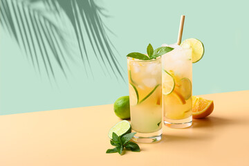 Wall Mural - Two summer cold mojito cocktails with orange and lime slices on green and beige background. Refreshing summer beverage with sunny shadow.