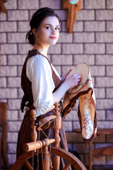 Portrait of Young Lovely Caucasian Brunette Woman Posing With Fancywork Hoop in Front of Spinning Wheel in Retro Dress In Rural Environment.