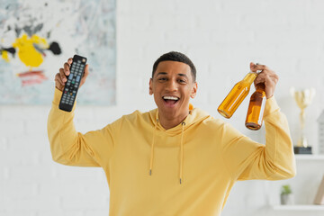 Wall Mural - smiling african american man with TV remote and bottles of beer in celebrating pose on blurred background