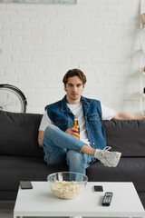 Wall Mural - young thoughtful man sitting with beer on couch near table with popcorn at home