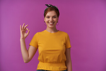 Wall Mural - Portrait of attractive european young brown eyed woman looking at you wearing stylish summer yellow t-shirt and black bandana emotion happy glad okay fingers posing isolated against purple background