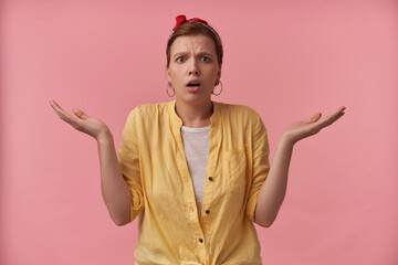 Wall Mural - Portrait of attractive young confused brown eyed european woman 20s with natural makeup wearing yellow shirt and red bandana posing against pink wall emotion anxiety astonishment with arms asides