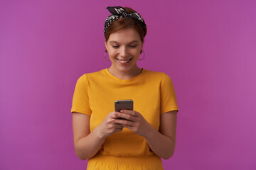 Wall Mural - Portrait of attractive young woman natural makeup wearing stylish trendy summer clothes and bandana with arms hold the phone emotion reading looking down smiling happy face posing against purple wall