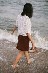 Wall Mural - Carefree hipster woman walking barefoot on sandy beach and sea waves in the evening, enjoying calm moment. Casual young female with flip flops in hand relaxing on seashore