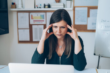 Wall Mural - Young tired female online professor having headache.
Tired woman sitting in her home office during online class break and holding her head with hands tired of computer screen.