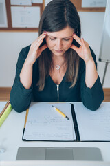 Wall Mural - Tired female online professor having headache.
High angle view of woman teacher sitting in her home office desk during online class break and holding her head with hands tired of computer screen.