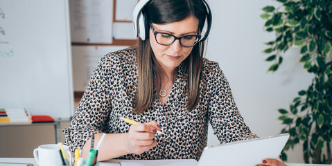 Wall Mural - Female teacher or student with headset holding tablet and writing notes in papers. Close up of experienced professor holding online class for e-learning student by video call online from home office.