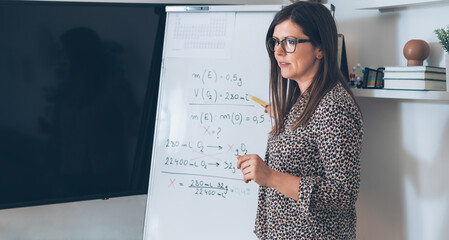Wall Mural - Teacher recording video tutorial for e-learning class.
Female professor standing and explaining the assignment pointing on the whiteboard and using camera for live streaming online on the internet.