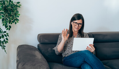 Wall Mural - Smiling Businesswoman Say Hello to Teamwork Colleague in Video Conference Meeting while Sitting on Couch at Home.
Happy business woman with glasses looking at digital tablet and waving with hands.