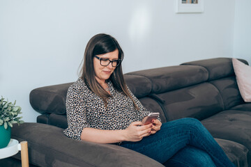 Wall Mural - Cheerful Woman Sitting on Sofa and Using Mobile Phone at Home
Smiling businesswoman working at home, calling or typing message on smartphone next to the plants in flowerpot.