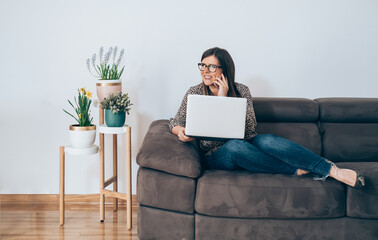 Wall Mural - Happy Business Woman Sitting on Sofa Talking on Mobile Phone and Holding Laptop
Cheerful smiling businesswoman relaxing on couch in living room at home and calling somebody on smartphone.