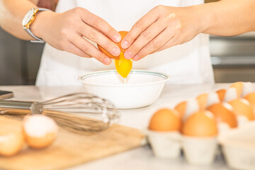 Young chef in white apron crack fresh egg, yolk dropping in a bowl. Peparing raw ingredient for making healthy cuisine, depth of field. Hands holding eggshell with blurred cooking items. Homemade meal