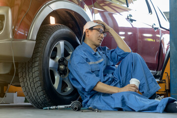 Garage worker or technician sit and lean on car wheel and he look tired or unhappy in workplace.