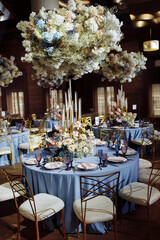 Round dining tables, covered in d blue cloth, served with porcelain and blue and purple glasses and richly decorated with flowers, flowers under the tables. Bokeh gold candles on