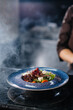 A professional chef serves a freshly prepared salad of tomato and veal greens under a glass hood with thick smoke. Beautiful smoky serving in the restaurant.