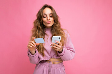 Beautiful young blonde curly woman wearing pink clothes isolated over pink background using mobile phone paying online shopping through credit card looking at smartphone display