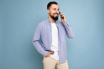 Side-profile Photo shot of smiling good looking young brunet bearded man wearing casual blue shirt and white t-shirt poising isolated on blue background with empty space holding in hand and