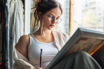 Wall Mural - Portrait of a pretty female artist painting on canvas in her art studio sitting next to the window. A creative woman painter with eyeglasses painting with oil searching for imagination in the workshop