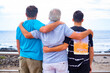 Family group, three males, father, adult son and teen grandson embracing each other looking at horizon over water