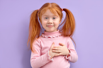 Caucasian little girl looking at camera and holding hands on heart gesture of love. Studio portrait of thankfull young girl with happy facial expression