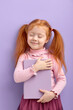 © alfa27 - red haired little girl isolated over purple background. sweet little girl in casual wear hug book, with eyes closed. school, education concept.