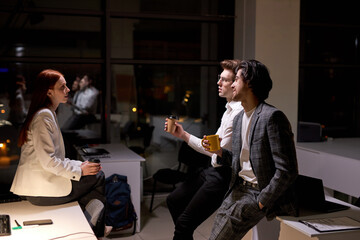 Side view on nice young office workers, colleagues having talk after hard working day, at night. Redhead woman and two guys in formal wear discussing, taking a break, bussiness team in boardroom